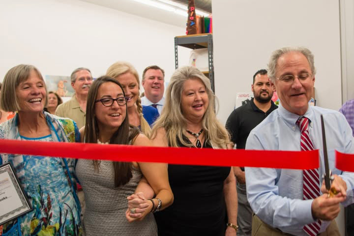 A man cuts a red ribbon as several people watch excitedly