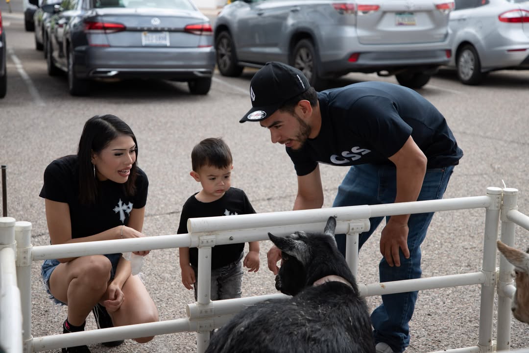 A mom and dad squat next to their young son as he pets a goat