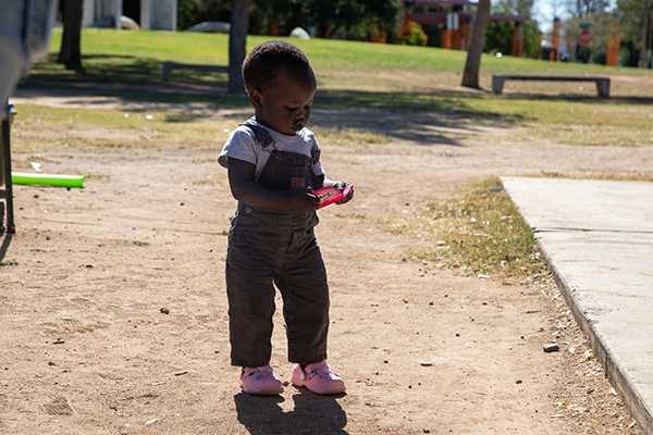 A toddler in brown overalls and pink Crocs holds a pack of gum