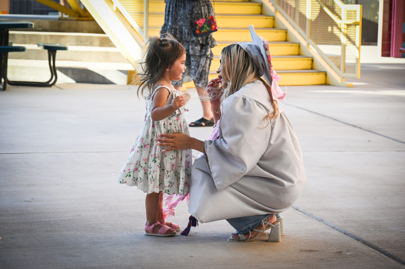 A TAP student kneels down in her cap and gown to talk to her young daughter