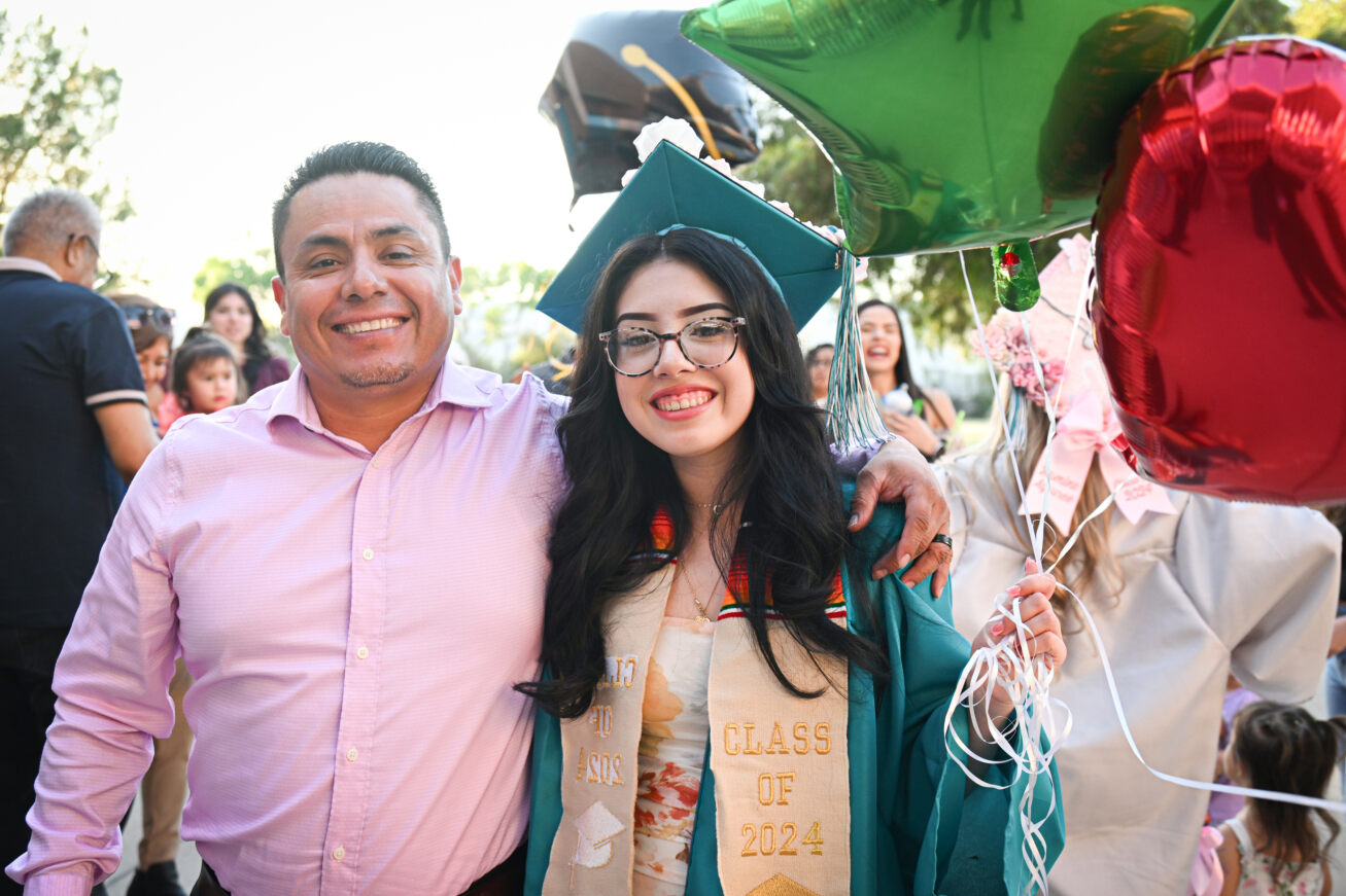 A TAP student smiles in her cap and gown holding balloons next to her father